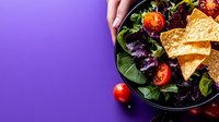 Fresh garden salad with crispy tortilla chips, cherry tomatoes and purple basil leaves served in black bowl on vibrant purple background with copy space.