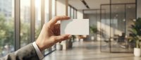 Professional businessman holding blank white business card in modern office lobby with glass walls and natural lighting for corporate branding mockup.