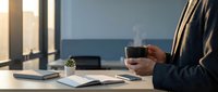 Professional businessman holding steaming coffee cup at modern office desk with laptop and documents during morning work routine.