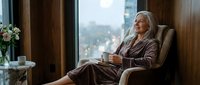 Elegant senior woman with gray hair relaxing in comfortable chair by window, enjoying peaceful morning coffee moment in modern home interior.