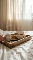 Wooden tray with steaming coffee, croissant, and welcome card on a hotel bed with soft morning light and sheer curtains.