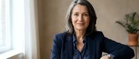 Mature woman with long dark hair and light skin tone wearing navy blazer over dark shirt, seated indoors with neutral beige wall background and potted plant, professional business portrait.