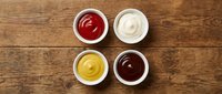 Four small white bowls with ketchup, mayonnaise, mustard, and barbecue sauce arranged on a rustic wooden table, top view.
