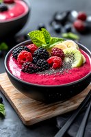 Pink smoothie bowl topped with fresh blackberries, raspberries, kiwi slices, banana, chia seeds and mint leaves served in black bowl on wooden board.