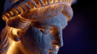 Close-up profile view of bronze Statue of Liberty head with dramatic lighting and dark background, showing intricate crown details and facial features in metallic texture.