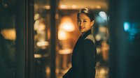 Professional Asian businesswoman in black blazer smiling confidently in modern office building lobby with warm ambient lighting during evening hours.
