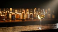 Glass of white wine on wooden bar counter with blurred whiskey bottles in background creating warm ambient lighting for restaurant and hospitality concepts.