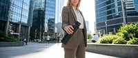 Middle-aged Caucasian woman in professional business attire standing in urban downtown setting surrounded by modern skyscrapers, wearing brown blazer and white shirt, suitable for corporate, finance, and business concepts.