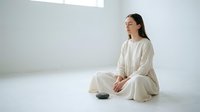 Young East Asian woman in casual beige linen clothing sitting cross-legged on white floor in minimalist interior space, suitable for wellness, lifestyle, and meditation content.
