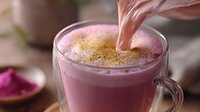 Pink beetroot latte with golden spice powder being sprinkled on top of frothy milk foam in glass mug, close up view of trendy healthy drink preparation.