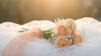 Elegant peach ranunculus wedding bouquet with baby's breath and flowing ribbon on white fabric in soft golden sunlight for bridal ceremonies and romantic events.