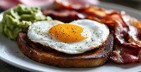 Sunny side up egg on toasted bread with crispy bacon strips and fresh avocado served on gray plate, close up view of breakfast meal with black pepper seasoning.