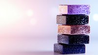Stack of glittery cosmetic eyeshadow blocks in purple, black, and pink tones on soft pastel background with bokeh effect.
