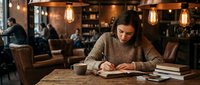 Woman with dark hair and olive complexion, wearing a striped shirt, reading a book at a wooden table in a warm, ambient-lit café or lounge with hanging Edison bulbs and leather seating.