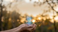 Glowing neon footprint symbol floating above open palm against blurred sunset forest background with bokeh lights, representing environmental awareness and sustainable living concepts.