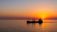 Silhouette of fishing boat with crew at golden hour sunrise over calm ocean waters creating peaceful seascape with warm orange and pink sky colors.