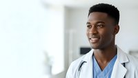 Young African American male doctor in white coat and blue scrubs smiling confidently in modern hospital setting for healthcare marketing materials.