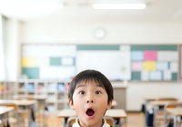 Young East Asian girl with dark hair and surprised expression in bright classroom with desks, chairs, and colorful bulletin board for educational content and learning materials.