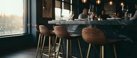 Modern bar interior with wooden stools at counter, warm pendant lighting, and large windows overlooking winter landscape. Contemporary hospitality design for restaurants and lounges.
