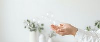 Hand holding soap bubbles with blurred green plants in vase background, representing cleanliness, hygiene, and wellness for skincare or cleaning product marketing.