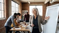 Mature woman with gray hair and light skin tone, wearing business casual attire, leads a startup meeting in a modern loft office. She gestures toward a whiteboard while diverse team members collaborate at a wooden table, ideal for corporate training and business content.