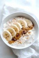 Fresh banana slices with peanut butter and dark chocolate shavings on creamy yogurt in white ceramic bowl, topped with cinnamon on white fabric background.