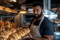 Professional African American male baker arranging fresh croissants on display shelves in commercial bakery kitchen wearing apron and smiling at camera.