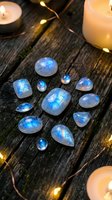 Moonstone gemstones with blue flash arranged on rustic wooden surface with glowing candles and fairy lights creating mystical atmosphere for jewelry design.