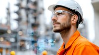 Caucasian male engineer in orange safety uniform and white hard hat with protective glasses at industrial facility examining digital interface.
