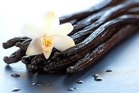 Aromatic vanilla pods with fresh white orchid flower and scattered seeds on blue surface, macro food photography with soft natural lighting and shallow depth.