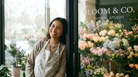 Young woman with dark hair and South Asian ethnicity, wearing a beige cardigan and cream lace top, smiling while standing in a modern flower shop doorway surrounded by potted plants and fresh floral arrangements in pink, white, and green tones.