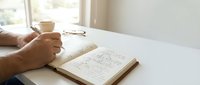 Male hands holding coffee cup while reviewing architectural floor plans and blueprints on wooden desk near bright window for home design and construction planning.