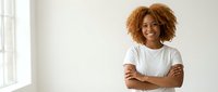 Happy young African American woman with curly hair smiling confidently with arms crossed in bright modern office space for business and career content.