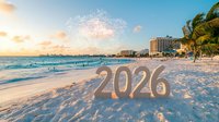 Festive 2026 numbers on tropical beach at sunset with fireworks, palm trees and hotels along coastline. Waves rolling onto white sand shore during golden hour.