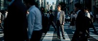 Young professional businessman in gray suit standing confidently in busy urban crosswalk during rush hour with blurred pedestrians and city skyline background.