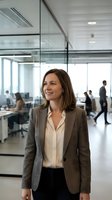 Middle-aged woman with brown hair wearing professional business attire including taupe blazer and cream blouse, smiling in modern glass-walled office environment with colleagues working in background.