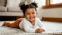 Happy mixed race toddler girl with curly hair in space buns lying on carpet at home, smiling joyfully during playtime in cozy living room interior.