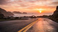 Coastal highway stretches toward golden sunset over ocean and mountains, with yellow center line marking road ahead. Perfect for travel, adventure, and transportation themes.