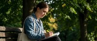 Young Asian woman writing in notebook while sitting on park bench surrounded by green trees during sunny day for journaling and creative writing.