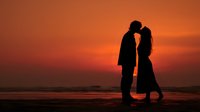 Romantic couple silhouette kissing at sunset on beach with orange sky and ocean waves creating intimate moment for love and relationship concepts.