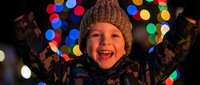 Happy young Caucasian boy in winter knit hat celebrating with arms raised against colorful Christmas lights bokeh background during holiday season festivities.