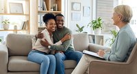Happy African American couple embracing on couch during therapy session with mature Caucasian counselor in bright modern office setting.