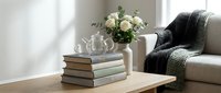 Cozy living room interior with stack of books, white roses in vase, glass teapot and knitted throw blanket on beige sofa creating warm home atmosphere.