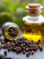 Black peppercorns scattered on dark surface with glass bottle of essential oil in background, macro food photography with selective focus and natural light.