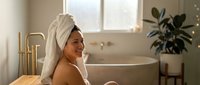 Young woman relaxing in modern bathroom after bath with towel wrapped around head, enjoying peaceful spa-like wellness routine at home.