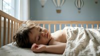 Peaceful Asian toddler sleeping soundly in wooden crib with hot air balloon wall decals in bright nursery room for childcare and parenting concepts.