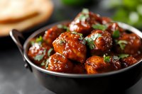 Glazed chicken meatballs in sweet and spicy Asian sauce garnished with fresh herbs, served in black ceramic bowl on dark background. Perfect for menu design.