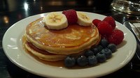 Stack of golden pancakes topped with maple syrup, fresh banana slices, raspberries and blueberries served on white plate, close up view of breakfast food.