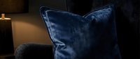 Luxurious deep blue velvet cushion with elegant piping detail displayed on modern furniture in warm ambient lighting, perfect for home decor and interior design projects.