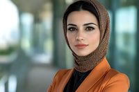 Professional Middle Eastern woman wearing hijab and orange blazer poses confidently in modern office setting with soft natural lighting and blurred background.